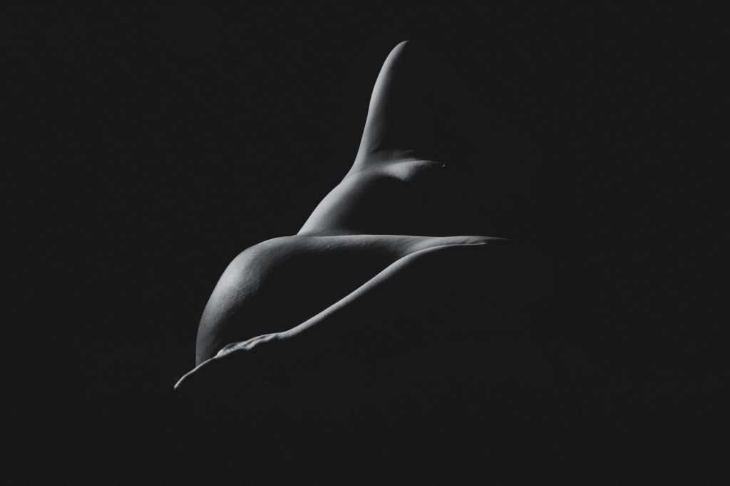 A little light falling on the naked women in the dark, showcasing her side breast and foot. Featured in the blog post "Boudoir Inspo: Nudescape or Bodyscape Photography" nudescape, beautiful boudoir, boudoir style, imagery style, Colorado boudoir photographer, Body positive boudoir, Body Image, inspiration, style, black and white imagery, composition. Imagery by Embodied Art Boudoir.