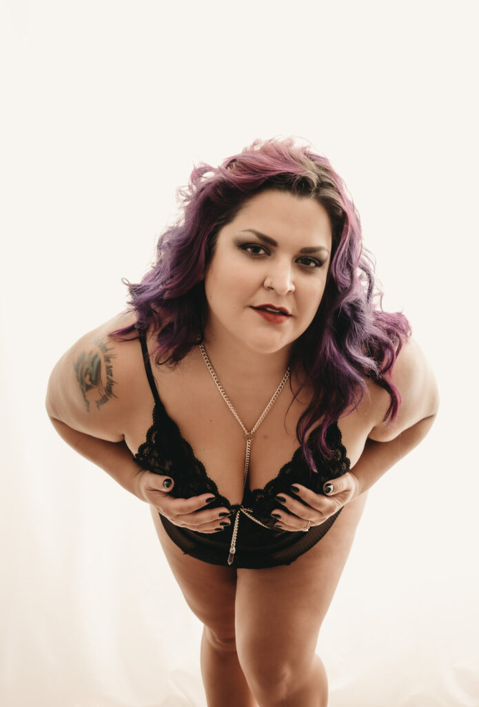 pretty older plus-size Woman with dyed purple hair leaning forward wearing a black body suit. Featured in the Blog Boudoir Outfit Ideas & Inspiration for 2024: How to Pick the Perfect Boudoir Look. Boudoir photography, colorado boudoir, denver boudoir, boulder boudoir, colorado springs boudoir, boudoir ideas, boudoir poses, boudoir inspiration, boudoir outfits, curvy boudoir, plus size lingerie, lingerie, photoshoot outfits, flattering outfits, curvy lingerie, lingerie look, lingerielook photoshoot, lingerie outfit, lingerie outfit bedroom, lingerie party, outfit ideas. image by Embodied Art Boudoir. 