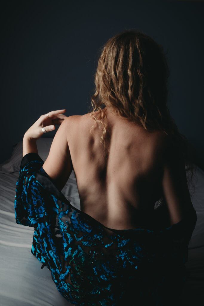 Woman with curly blond hair back exposed, wearing only a blue robe. Featured in the Blog Boudoir Outfit Ideas & Inspiration for 2024: How to Pick the Perfect Boudoir Look. Boudoir photography, colorado boudoir, denver boudoir, boulder boudoir, colorado springs boudoir, boudoir ideas, boudoir poses, boudoir inspiration, boudoir outfits, curvy boudoir, plus size lingerie, lingerie, photoshoot outfits, flattering outfits, curvy lingerie, lingerie look, lingerielook photoshoot, lingerie outfit, lingerie outfit bedroom, lingerie party, outfit ideas. image by Embodied Art Boudoir. 
