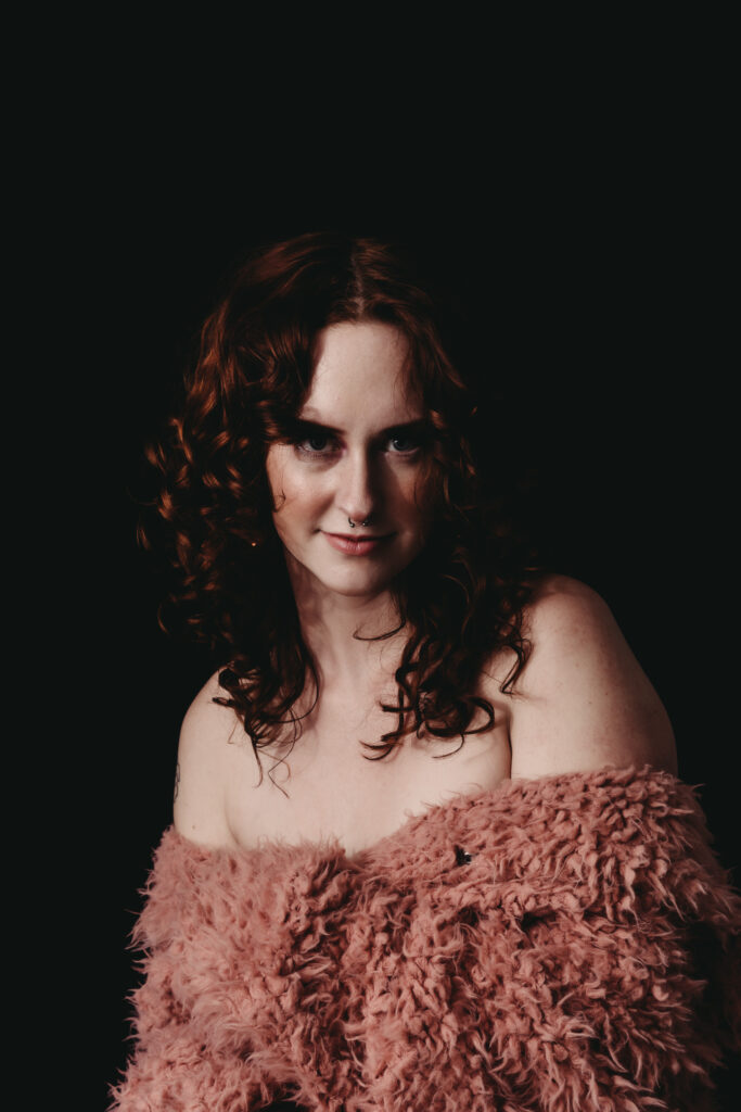 Red haired young Woman sitting facing the camera, wearing a fluffy pink sweater and curly hair. Featured in the Blog Boudoir Outfit Ideas & Inspiration for 2024: How to Pick the Perfect Boudoir Look. Boudoir photography, colorado boudoir, denver boudoir, boulder boudoir, colorado springs boudoir, boudoir ideas, boudoir poses, boudoir inspiration, boudoir outfits, curvy boudoir, plus size lingerie, lingerie, photoshoot outfits, flattering outfits, curvy lingerie, lingerie look, lingerielook photoshoot, lingerie outfit, lingerie outfit bedroom, lingerie party, outfit ideas. Photography by Embodied Art Boudoir. 