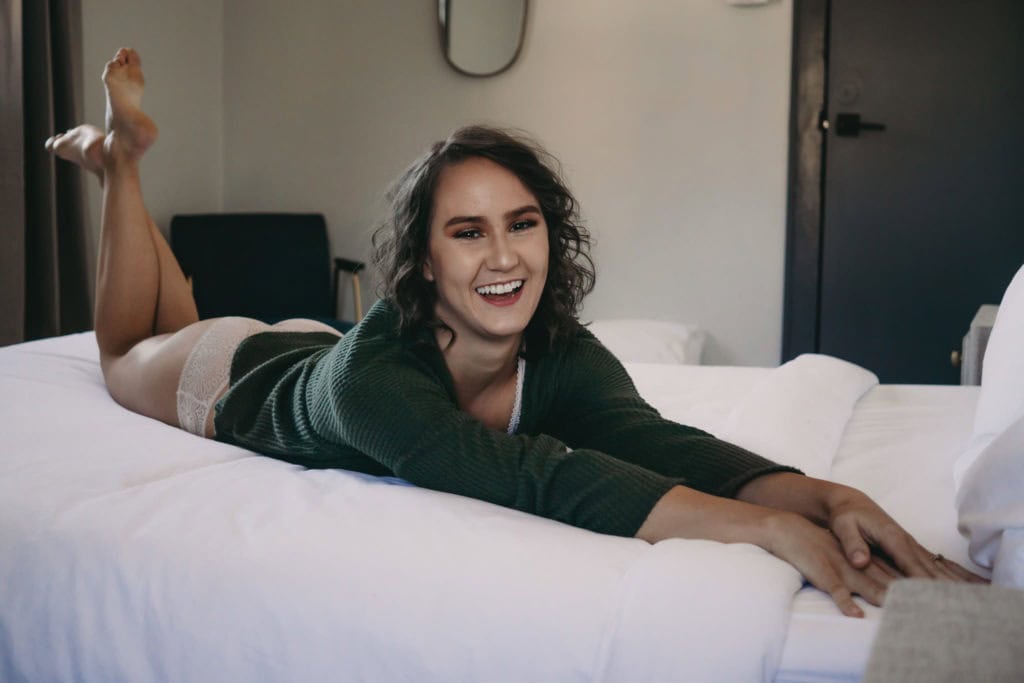 young brunette woman with short curly hair smiling with crossed legs lying on a bed, wearing a dark green cardigan and white covered panties. Featured in the blog "You Don't Have To Be Naked (Or Even Show A Ton Of Skin) For A Boudoir Photoshoot!" Modest photography, modest boudoir, modest photoshoot outfit, modest outfit ideas, boudoir photography, boudoir photoshoot, boudoir session, covered up, Colorado photographer, Denver boudoir photographer, Colorado boudoir photography, studio photography, body positivity, body positive photography. Photo by Embodied Art Boudoir.