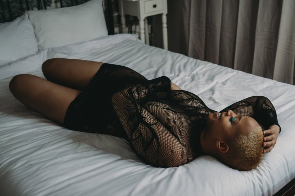 Black woman with a short haircut that is dyed blonde with bright blue eyeliner, wearing a long sleeves bodysuit lounging on a bed with crispy white sheets smiling joyfully. Featured in the blog "You Don't Have To Be Naked (Or Even Show A Ton Of Skin) For A Boudoir Photoshoot!" Modest photography, modest boudoir, modest photoshoot outfit, modest outfit ideas, boudoir photography, boudoir photoshoot, boudoir session, covered up, Colorado photographer, Denver boudoir photographer, Colorado boudoir photography, studio photography, body positivity, body positive photography. Image by Embodied Art Boudoir.