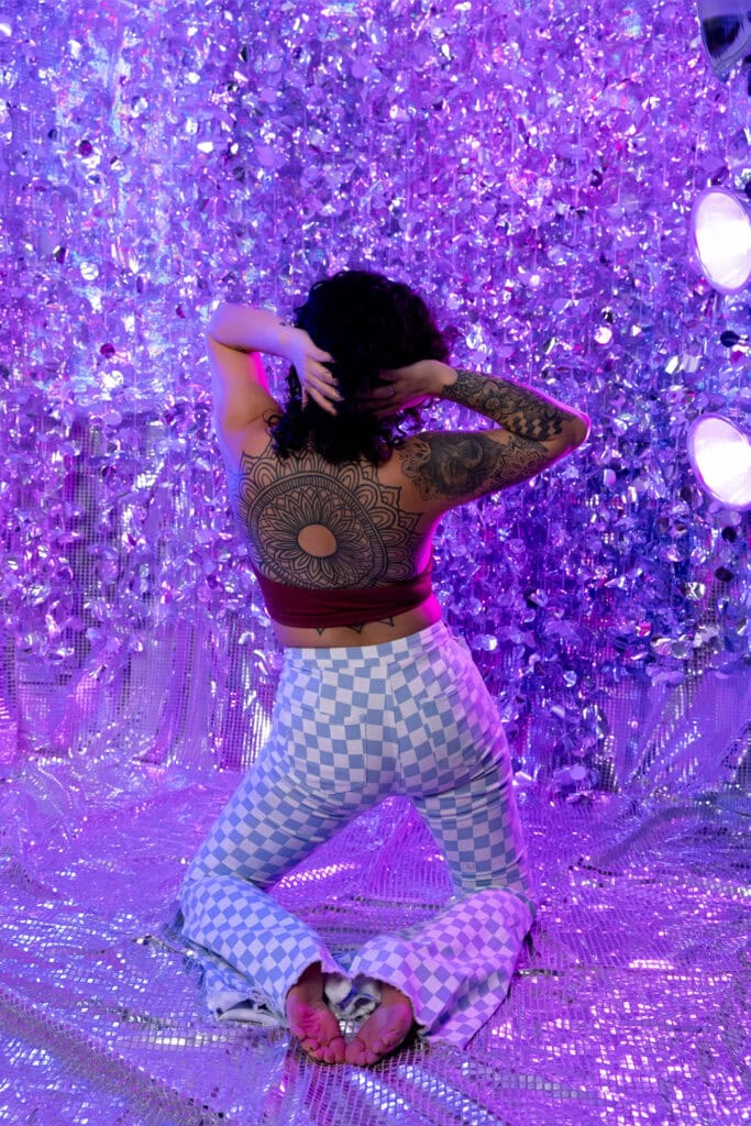 Woman shirt short curly black hair posing in front of a disco ball background wearing checkered blue and white jeans with a dark red halter top. Featured in the blog "You Don't Have To Be Naked (Or Even Show A Ton Of Skin) For A Boudoir Photoshoot!" Modest photography, modest boudoir, modest photoshoot outfit, modest outfit ideas, boudoir photography, boudoir photoshoot, boudoir session, covered up, Colorado photographer, Denver boudoir photographer, Colorado boudoir photography, studio photography, body positivity, body positive photography. Picture by Embodied Art Boudoir.