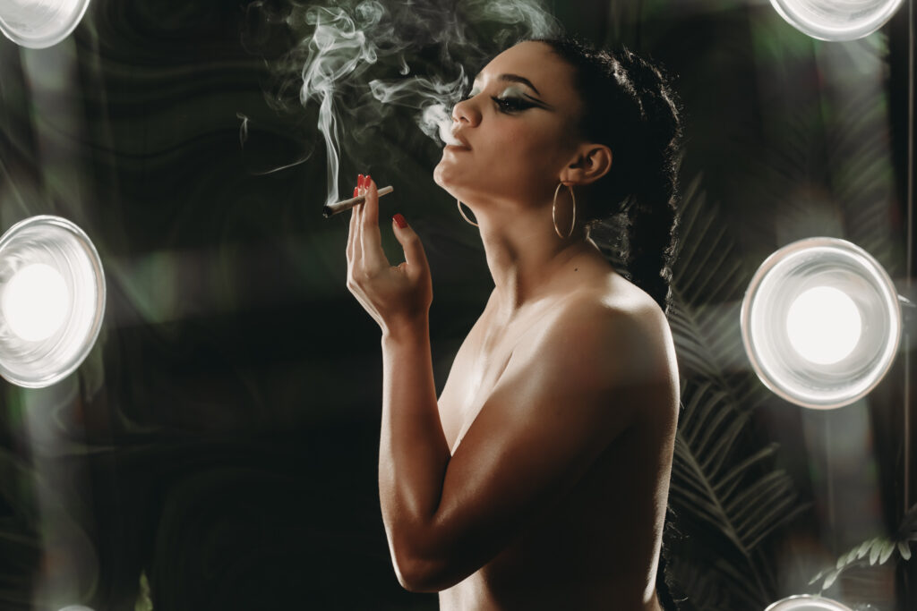 Beautiful brunette woman with a tight braid pony tail smoking a joint with the smoke puffing dramaticaly in the air insid the glam room with spotlights on her. Featured in the blog "Outfit & Prop Ideas For The Green Room: Boudoir 420 Photoshoot." boudoir, sensual, sexy, playful, flirty, provocative, daring, confident, edgy, creative, unique, expressive, intimate, vibrant, lush, dramatic, whimsical, mysterious, artistic, stylish, bold, fun, liberating, empowering, seductive, luxurious, unapologetic, radiant, magnetic, alluring, captivating, enticing, glamorous, seductive, spirited, mischievous, charismatic, empowering, personal, transformative, celebratory, indulgent, immersive, photogenic. Picture by Embodied Art Boudoir