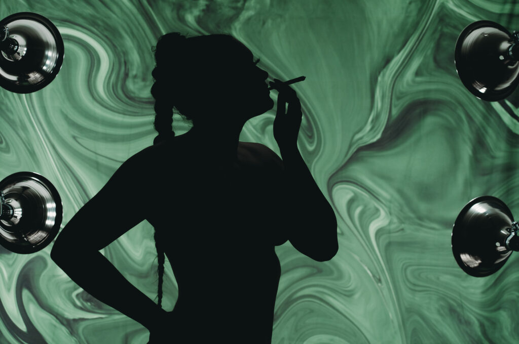 Womans silhouette of braid and her back as she lights and smokes from a joint in front of a trippy and dreamy green background. . Featured in the blog "Outfit & Prop Ideas For The Green Room: Boudoir 420 Photoshoot." boudoir, sensual, sexy, playful, flirty, provocative, daring, confident, edgy, creative, unique, expressive, intimate, vibrant, lush, dramatic, whimsical, mysterious, artistic, stylish, bold, fun, liberating, empowering, seductive, luxurious, unapologetic, radiant, magnetic, alluring, captivating, enticing, glamorous, seductive, spirited, mischievous, charismatic, empowering, personal, transformative, celebratory, indulgent, immersive, photogenic. Pic by Embodied Art Boudoir