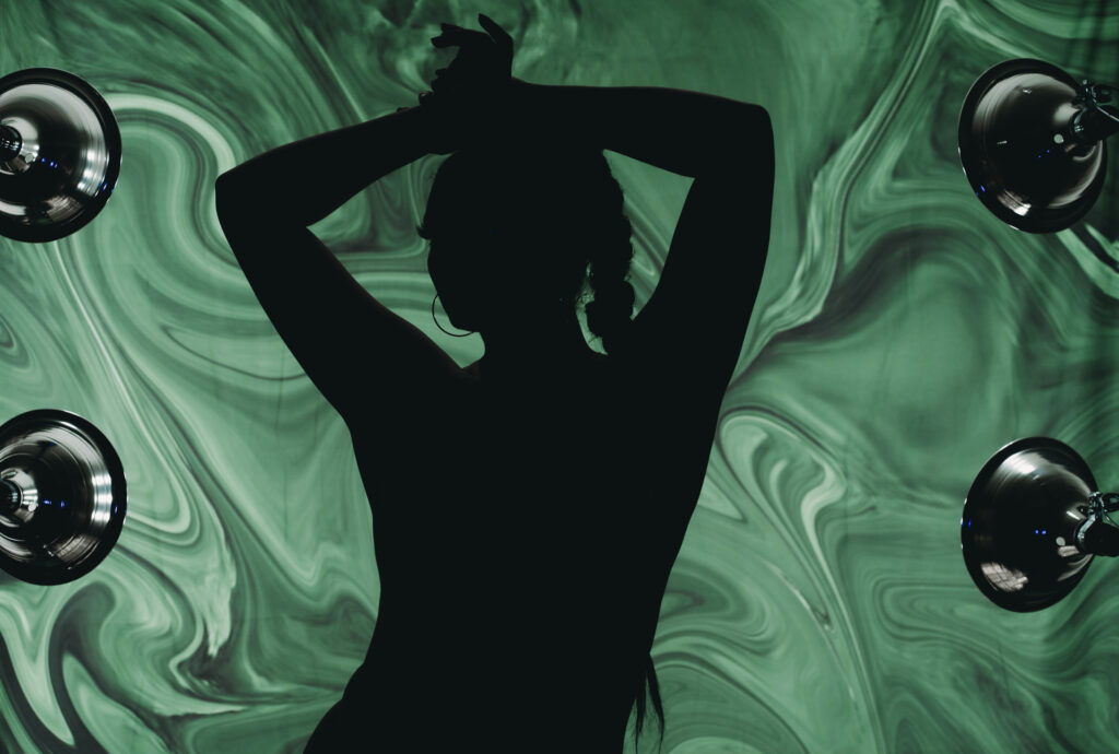 Silhoutte of a woman posing with her hands behind her head featuring her eyelashes, braid in front of a trippy green background. . Featured in the blog "Outfit & Prop Ideas For The Green Room: Boudoir 420 Photoshoot." boudoir, sensual, sexy, playful, flirty, provocative, daring, confident, edgy, creative, unique, expressive, intimate, vibrant, lush, dramatic, whimsical, mysterious, artistic, stylish, bold, fun, liberating, empowering, seductive, luxurious, unapologetic, radiant, magnetic, alluring, captivating, enticing, glamorous, seductive, spirited, mischievous, charismatic, empowering, personal, transformative, celebratory, indulgent, immersive, photogenic. Image by Embodied Art Boudoir