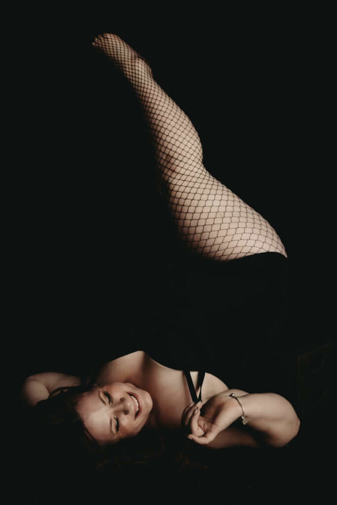 Woman lying down with legs in the air playfully smiling. Wearing a black body suit and fishnet stockings. Photo by Embodied Art Boudoir. Boudoir photography, self-care, journal, journal inspiration, journaling, journal addict, journal lover, journal inspo, self-care tips, self-care first, self-care routine, self-care tips, self-care daily, healthy choices, healthy habits, habits, success habits, habits for success, me time, quotes to live by, colorado, colorado photographer, Denver boudoir, Denver boudior, Colorado boudior, Embodied art boudoir, boudior, boudoir, boudoir photo, photographer