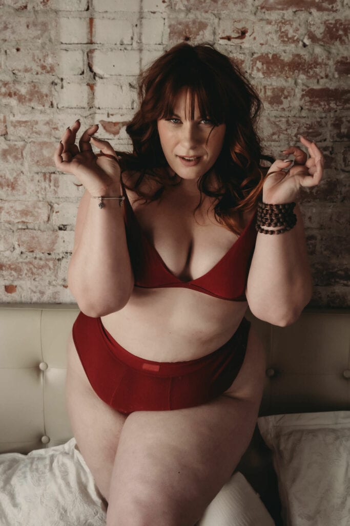 Plus size red-haired woman with curly hair and bands standing confidently in front of a white and red brick wall looking sultry wearing a matching red bra and briefs with brown chunky bracelets on her wrist. Featured in the blog, "How To Improve Your Body Image." . Denver boudoir photographer, denver boudoir photography, boudoir, colorado boudoir, denver boudoir, body positive, body positivity, body neutrality, body neutral, fat positive, plus size, petite, body image, body love, self love, self compassion, mirror work, boudoir photos, sexy photos, mindset, beautiful bodies, healing body image. Photo by Embodied Art Boudoir.