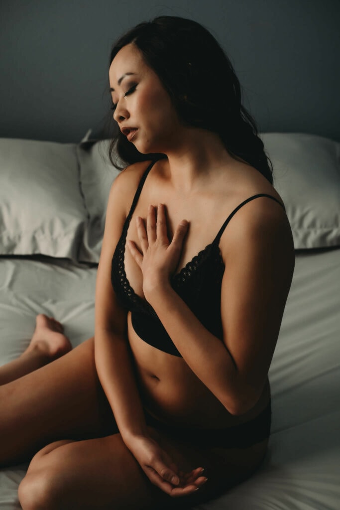 Woman with brunette curly hair sitting on bed, with her hand on heart and eyes closed wearing a matching black bralette and panties.Featured in the blog, "How To Improve Your Body Image." . Denver boudoir photographer, denver boudoir photography, boudoir, colorado boudoir, denver boudoir, body positive, body positivity, body neutrality, body neutral, fat positive, plus size, petite, body image, body love, self love, self compassion, mirror work, boudoir photos, sexy photos, mindset, beautiful bodies, healing body image. Image by Embodied Art Boudoir.