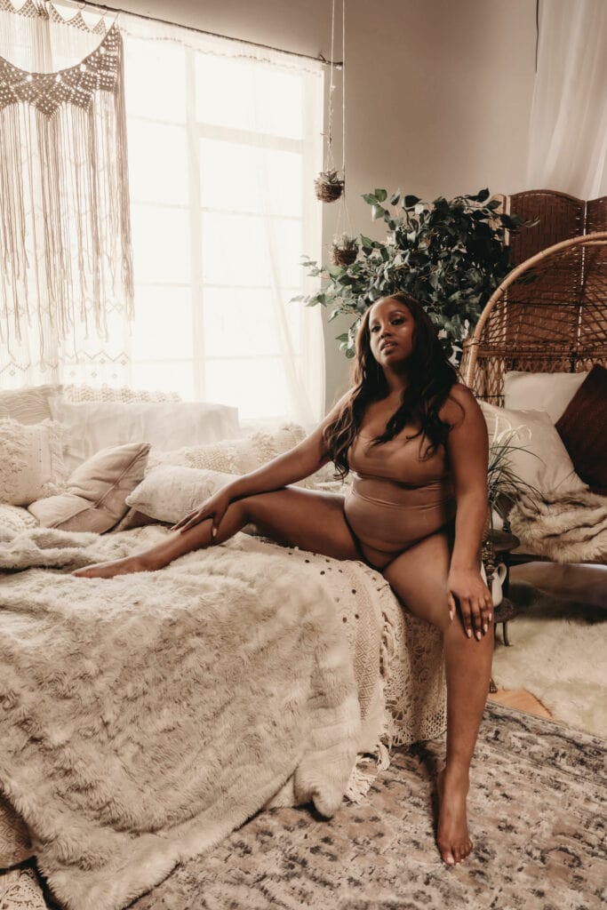 Black woman sitting on a furry blanket in a neutral bohemian style bedroom with plants, wearing a brown body suit. . Featured in the blog, "How To Improve Your Body Image." . Denver boudoir photographer, denver boudoir photography, boudoir, colorado boudoir, denver boudoir, body positive, body positivity, body neutrality, body neutral, fat positive, plus size, petite, body image, body love, self love, self compassion, mirror work, boudoir photos, sexy photos, mindset, beautiful bodies, healing body image. Photo by Embodied Art Boudoir.