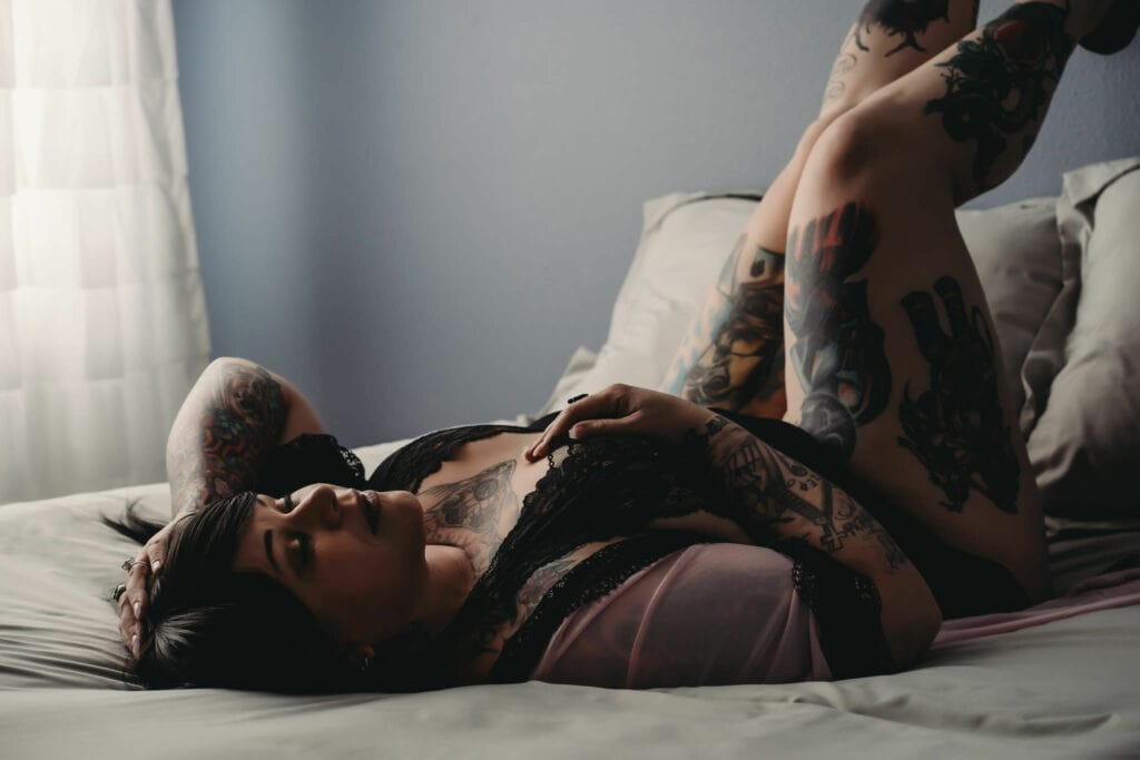 Plus-sized tattooed woman lying on her back on the bed with her hand on her chest, wearing a lacy black bralette and showcasing her many tattoos. Featured in the blog, "How To Improve Your Body Image." . Denver boudoir photographer, denver boudoir photography, boudoir, colorado boudoir, denver boudoir, body positive, body positivity, body neutrality, body neutral, fat positive, plus size, petite, body image, body love, self love, self compassion, mirror work, boudoir photos, sexy photos, mindset, beautiful bodies, healing body image. Picture by Embodied Art Boudoir.