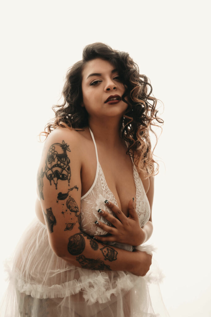 Woman with curly hair standing, with a sultry look on her face, holding her arms around her chest wearing a white see-through neglige. Photo by Embodied Art Boudoir. Intentions, intention setting, goal setting, boudoir photoshoot, boudior, buodoir, Denver boudoir, Colorado boudoir, boudoir photographer, self love, growth journey, affirmations, positive intentions, positive affirmations, self love journey, boudoir photography, Denver portrait photography, Denver portrait photographer, Boulder boudoir, fort collins boudoir, colorado springs boudoir, how to set an intention