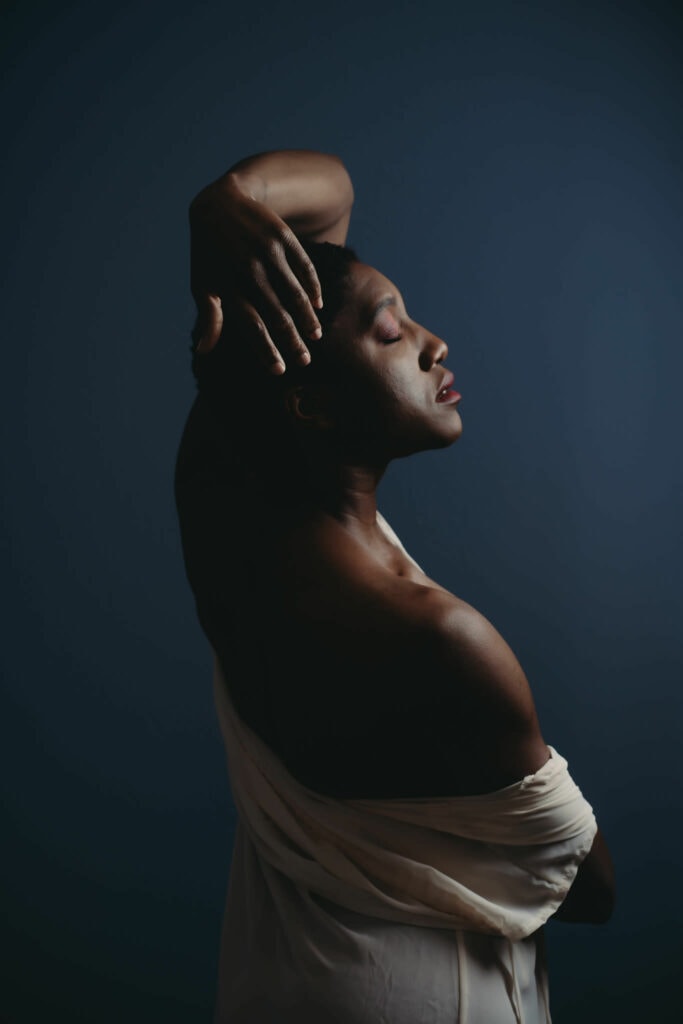 Black Woman with short hair standing with arm on head, eyes closed wearing a white robe draped over one shoulder. She has pink eyeshadow and red lipstick. Featured in the blog "Modest Boudoir Photography"Modest boudoir photography, Modest Fashion, Modest Woman, Fashion Trends, Shy, classy photography, classic boudoir, classy boudoir, feminine photography, colorado boudoir, denver boudoir, boulder boudoir, colorado springs boudoir, boudoir ideas, boudoir poses, boudoir inspiration, photography inspiration, boudoir session, boudoir photos. Image by Embodied Art Boudoir.