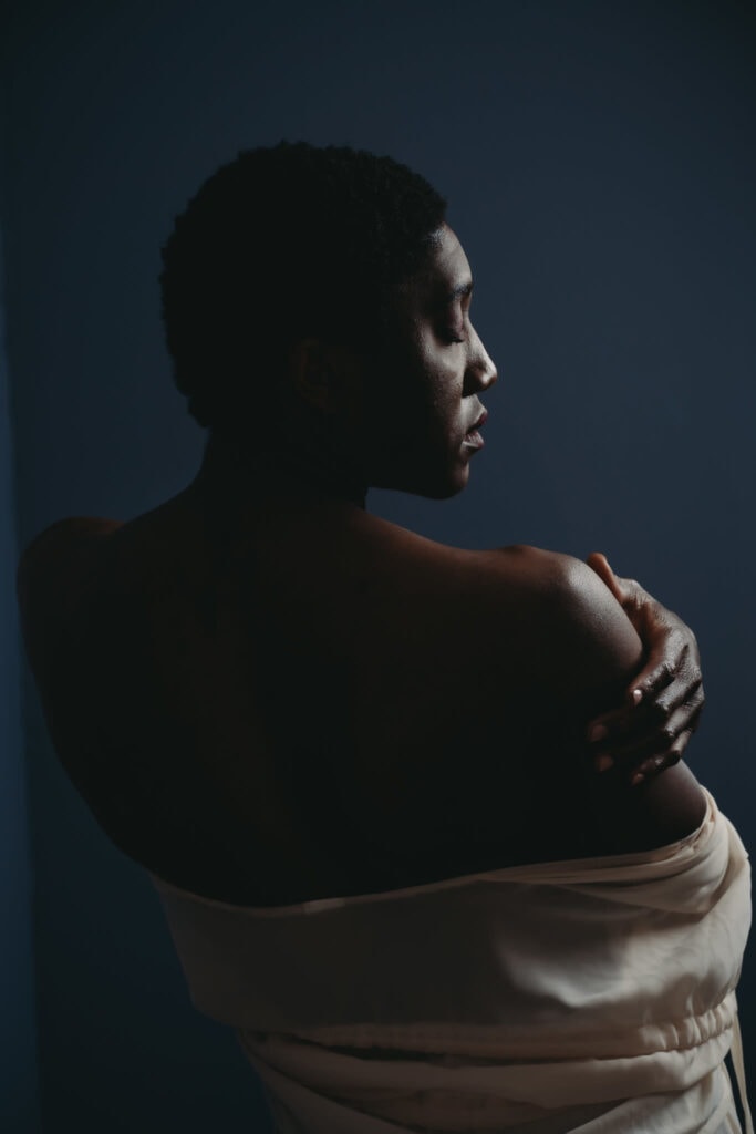 Black woman standing in front of a dark background exposing her right shoulder and wearing a white robe, showcasing her glowing skin from the side. Featured in the blog post "Dark & Moody Boudoir Photography Style"Dark and moody, black and white photography, intense images, high intensity, memorable, sensual photography, sensual boudoir, colorado boudoir, denver boudoir, boulder boudoir, colorado springs boudoir, boudoir ideas, boudoir poses, boudoir inspiration, photography inspiration, romantic boudoir, bridal boudoir. Imagery by Embodied Art Boudoir.