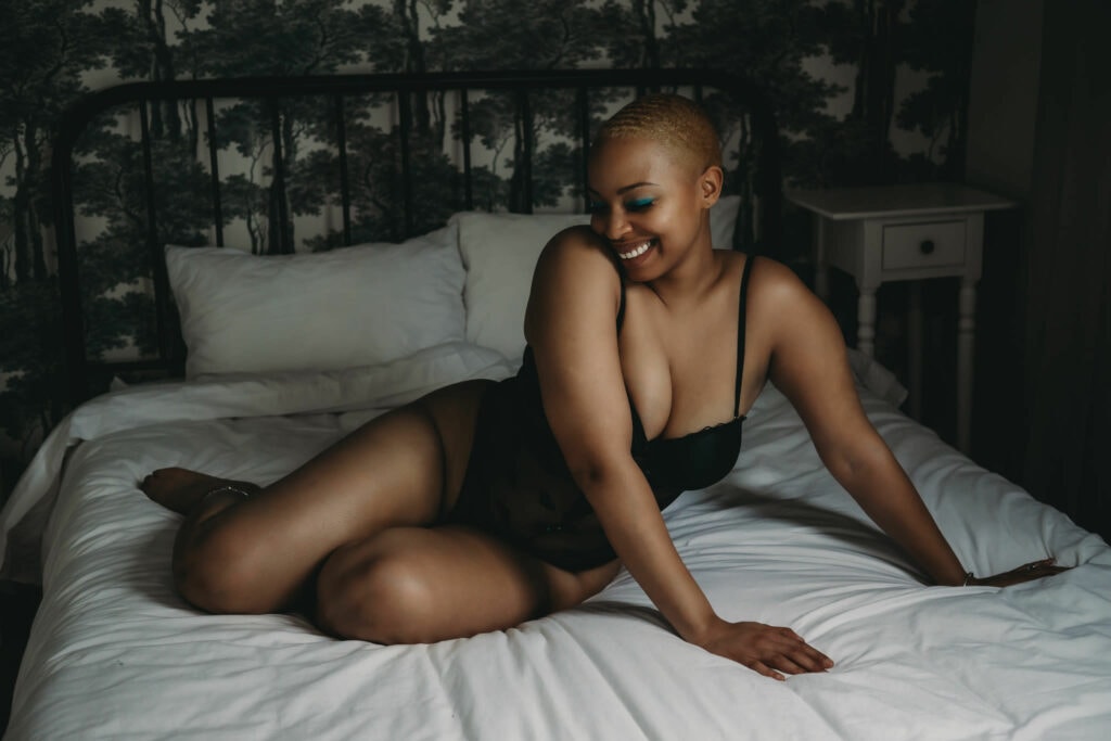 Young black Woman with very short hair lying on bed, smiling big showcasing her white teeth, wearing a black bodysuit and dark blue eyeshadow on her eyes. Featured in the blog post "Dark & Moody Boudoir Photography Style"Dark and moody, black and white photography, intense images, high intensity, memorable, sensual photography, sensual boudoir, colorado boudoir, denver boudoir, boulder boudoir, colorado springs boudoir, boudoir ideas, boudoir poses, boudoir inspiration, photography inspiration, romantic boudoir, bridal boudoir. Imagery by Embodied Art Boudoir.