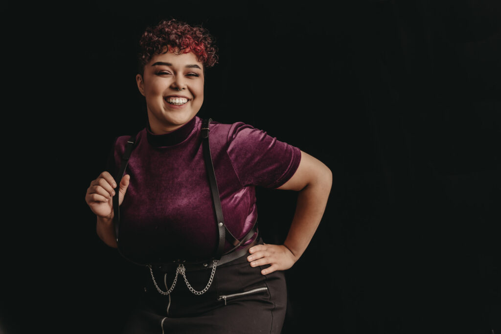 Woman standing facing the camera smiling at the camera, wearing a purple velvet shirt and black suspenders. Photo by Embodied Art Boudoir. outfit, outfit inspiration, look good, dress well, lingerie, casual, business casual, business formal, categories, clients, look good and feel good, stylish, personal style