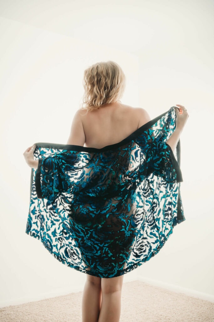 Curly hair blonde Woman standing up, wearing a blue opaque scarf and nothing underneath, showcasing her bare back. Featured in the blog "High Key Boudoir Photography Style". high key photography, high key, bright photohraphy, sensual photography, sensual boudoir, colorado boudoir, denver boudoir, boulder boudoir, colorado springs boudoir, boudoir ideas, boudoir poses, boudoir inspiration, photography inspiration, romantic boudoir, bridal boudoir, female empowerment, female photographer. Photography by Embodied Art Boudoir.