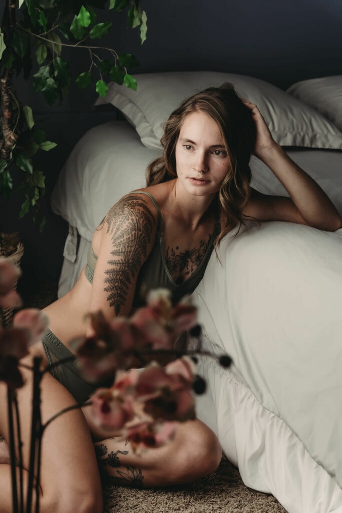 Young brunette Woman posing next to bed with crispy grey sheets, wearing green matching underwear, her leaf arm tattoo is on display and a fresh pink plant is in the foreground. Featured in the blog "Ethical Lingerie" Ethical shopping, ethical lingerie, sustainability, empowerment, shop sustainable, vote with your money, environmentally conscious, a living wage, lingerie piece, beautiful lingerie, shopping inspiration, mindfulness, boudoir lingerie, shop well, colorado boudoir, denver boudoir, boulder boudoir, colorado springs boudoir, boudoir ideas. Image by Embodied Art Boudoir