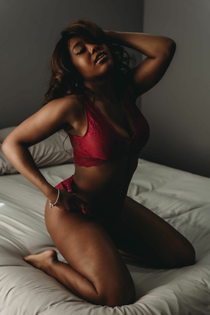 Black woman with curly brown hair kneeling on bed wearing a matching deep red bralette and panties. She is posing confidently, with one hand behind her head and other on hip. Featured in the blog "10 + Black & People of Color Owned Lingerie Brands You Need to Know About" black owned business, lingerie stores, people of color owned business, POC business, BIPOC business, successful females, business shout out, lingerie must haves, buy local, support your community, your money is a vote, black lives matter, colorado boudoir, denver boudoir, boulder boudoir, colorado springs boudoir, boudoir ideas, boudoir photography, photography inspiration. Image by Embodied Art Boudoir,