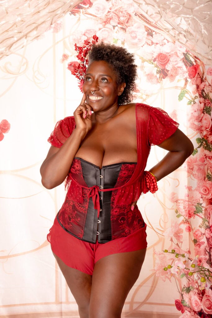 Mature black woman with short curly brown hair and natural lipstick posing in front of a colorful floral background wearing a black and red corset with matching red shorts. She poses confidently with a large smile on her face. Featured in the blog "10 + Black & People of Color Owned Lingerie Brands You Need to Know About" black owned business, lingerie stores, people of color owned business, POC business, BIPOC business, successful females, business shout out, lingerie must haves, buy local, support your community, your money is a vote, black lives matter, colorado boudoir, denver boudoir, boulder boudoir, colorado springs boudoir, boudoir ideas, boudoir photography, photography inspiration. Photography by Embodied Art Boudoir,