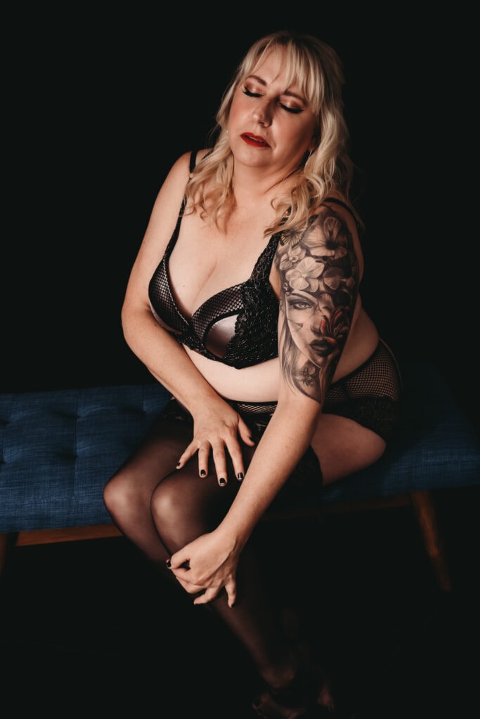 Blonde woman wearing a black bra with matching briefs and stockings, posing with her eyes closed and gold makeup sitting on a blue bench. Featured in the blog, "Can I Do a Boudoir Session If I Am Plus Size?" Curvy women, curvy girl style, plus size boudoir, plus size photography, plus sized women, fight the inner critic, self confidence, self love, love your body, stylish plus sized women, plus size lingerie, beautiful women, empowered women, photogenic, stunning boudoir images, boudoir imagery, boudoir picture, female photographer, denver boudoir, golden boudoir, golden colorado boudoir, boudoir inspiration. Photo by Embodied Art Boudoir.