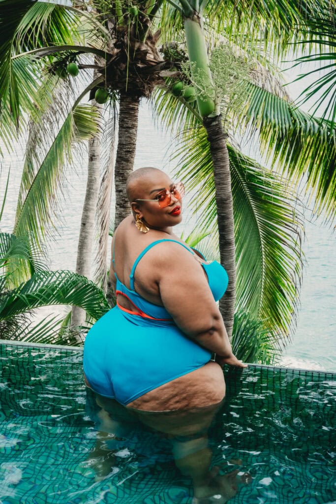 Plus sized black woman with a shaved head facing the camera froa in a pool with the ocean and gorgeous lush palm tree in the background, wearing a bright blue one piece, orange sunglasses and fun gold earrings. Featured in the blog, "Can I Do a Boudoir Session If I Am Plus Size?" Curvy women, curvy girl style, plus size boudoir, plus size photography, plus sized women, fight the inner critic, self confidence, self love, love your body, stylish plus sized women, plus size lingerie, beautiful women, empowered women, photogenic, stunning boudoir images, boudoir imagery, boudoir picture, female photographer, denver boudoir, golden boudoir, golden colorado boudoir, boudoir inspiration. Photography by Embodied Art Boudoir.