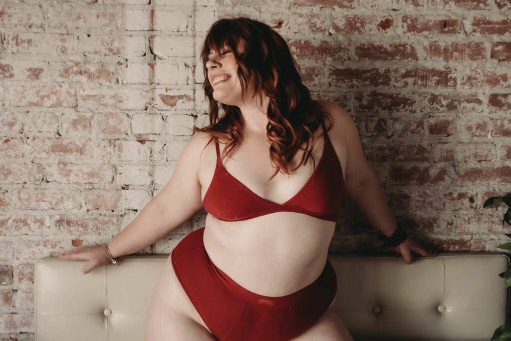 Plus size woman with red hair and bangs posing joyfully in front of a brick wall, wearing a red bralette and high-waisted red panties. Featured in the blog, "Can I Do a Boudoir Session If I Am Plus Size?" Curvy women, curvy girl style, plus size boudoir, plus size photography, plus sized women, fight the inner critic, self confidence, self love, love your body, stylish plus sized women, plus size lingerie, beautiful women, empowered women, photogenic, stunning boudoir images, boudoir imagery, boudoir picture, female photographer, denver boudoir, golden boudoir, golden colorado boudoir, boudoir inspiration. Imagery by Embodied Art Boudoir.