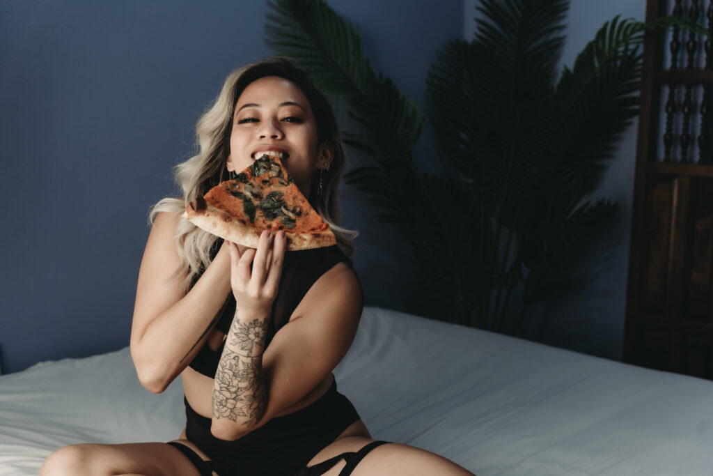 Blonde woman sitting on bed eating a slice of pizza in all black. Sexy silhouette of a woman in front of a green back ground. Photo by Embodied Art Boudoir. Golden, great location, savings, Colorado, female photographer, unique, savings, female photographer, boudoir photos