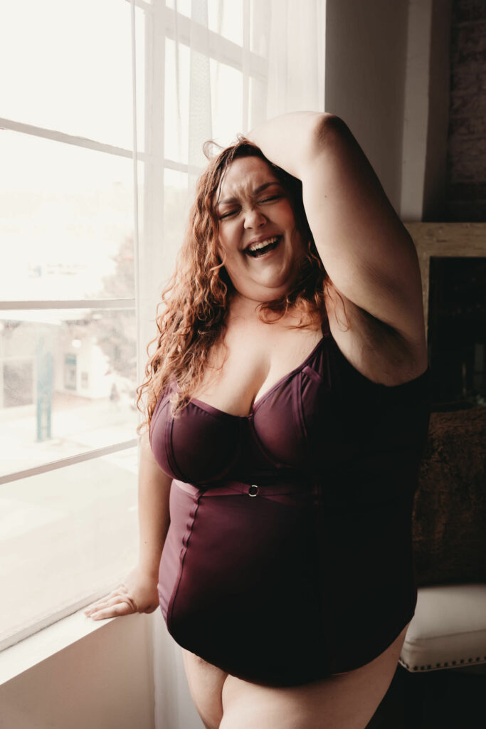 Woman standing next to window, wearing a deep purple body suit, smiling wide. Photo by Embodied Art Boudoir. Find Your photographer, boudoir photographer, boudoir photographers, right photographer, choose the right boudoir photographer, female boudoir photographer, boudoir images, classic boudoir, boudoir style, classy boudoir, sensual photography, sensual boudoir, colorado boudoir, denver boudoir, boulder boudoir, colorado springs boudoir, boudoir ideas, the perfect match.
