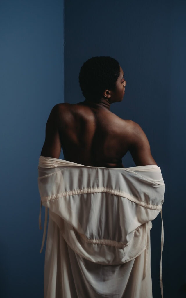 A black woman standing with back to camera, wearing a white robe draped over her sculpted back posing in front of a blue wall. Featured in the blog, "Best Accessories For Your Boudoir Photoshoot" Accessories, boudoir outfits, boudoir style, boudoir looks, accessorise, sheets, jewellery, scarf, sheet, creative outfits, sexy look, confidence, boudoir photoshoot, classy photography, sensual photography, sensual boudoir, colorado boudoir, denver boudoir, boulder boudoir, colorado springs boudoir, boudoir ideas. Imagery by Embodied Art Boudoir.