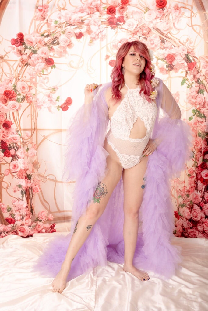 Woman with pink, curly hair posing in front of a floral pink and white background wearing a white body suit with a fluffy bright purple robe. Featured in the blog, "Best Accessories For Your Boudoir Photoshoot" Accessories, boudoir outfits, boudoir style, boudoir looks, accessorise, sheets, jewellery, scarf, sheet, creative outfits, sexy look, confidence, boudoir photoshoot, classy photography, sensual photography, sensual boudoir, colorado boudoir, denver boudoir, boulder boudoir, colorado springs boudoir, boudoir ideas. Photography by Embodied Art Boudoir.