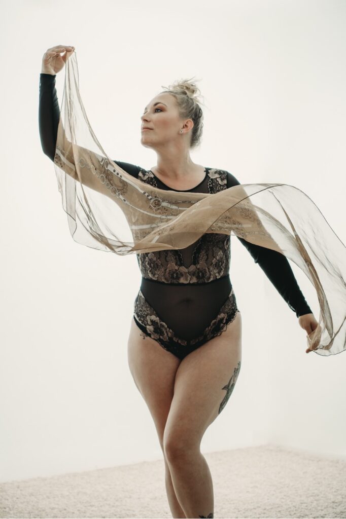 Blonde woman with a tight bun on her head standing in a dance pose, wearing a black body suit, and holding a white scarf floating beautifully. Featured in the blog, "Best Accessories For Your Boudoir Photoshoot" Accessories, boudoir outfits, boudoir style, boudoir looks, accessorise, sheets, jewellery, scarf, sheet, creative outfits, sexy look, confidence, boudoir photoshoot, classy photography, sensual photography, sensual boudoir, colorado boudoir, denver boudoir, boulder boudoir, colorado springs boudoir, boudoir ideas. Pic by Embodied Art Boudoir.