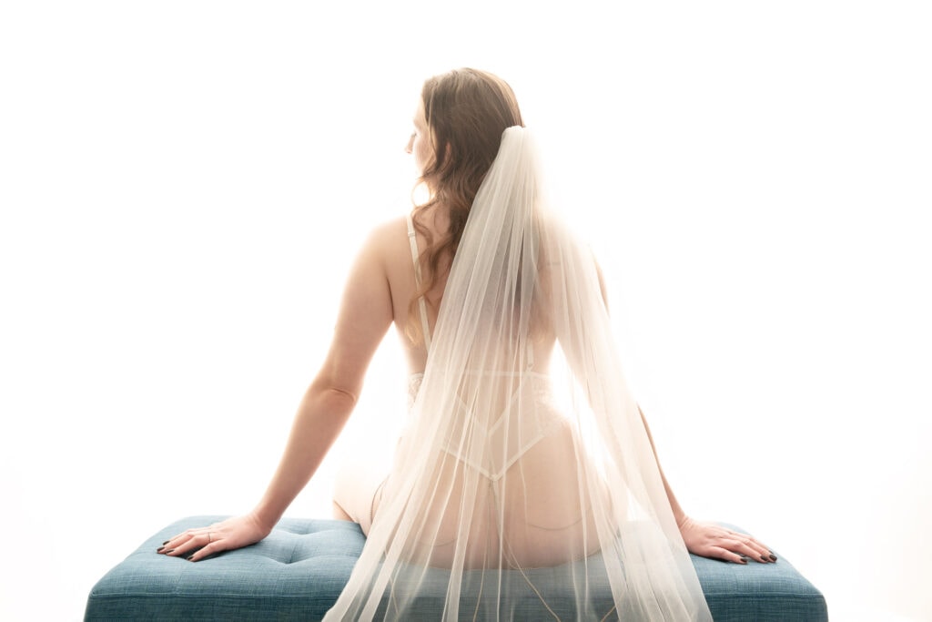 A brunette woman with long slightly curly hair sitting on a blue bench wearing a white g-string body suit with spaghetti straps and a long gorgeous veil, looking over her left shoulder. Featured in the blog, "Best Accessories For Your Boudoir Photoshoot" Accessories, boudoir outfits, boudoir style, boudoir looks, accessorise, sheets, jewellery, scarf, sheet, creative outfits, sexy look, confidence, boudoir photoshoot, classy photography, sensual photography, sensual boudoir, colorado boudoir, denver boudoir, boulder boudoir, colorado springs boudoir, boudoir ideas. Snapshot by Embodied Art Boudoir.