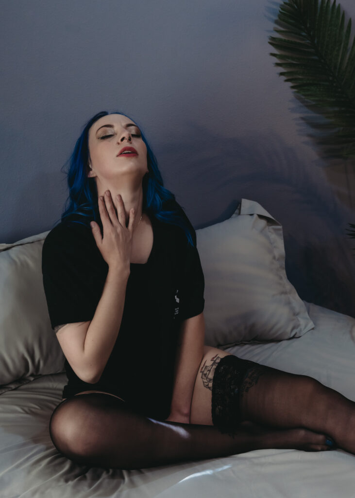 Woman with blue hair sitting on bed, touching her chest wearing a black t shirt and patnies and knee high socks. Featured in the blog "Non-Lingerie Boudoir Outfit Inspiration" non lingerie, outfit, bra, body suit, sexy looks, inspired, outfit inspiration, feel good, look good, wellness, boudoir inspo, female boudoir photography, colorado boudoir, denver boudoir, boulder boudoir, colorado springs boudoir, boudoir ideas, boudoir poses. Photo by Embodied Art Boudoir.