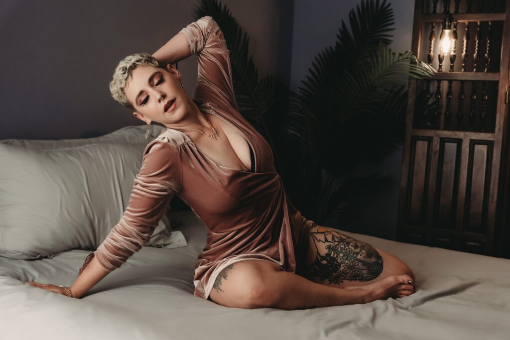 Blonde woman with a short pixie cut, wearing a velvet pink dress with a low cut and full sleeves, posing on a bed in a bedroom. Featured in the blog, "The Connection Between Mindfulness And Self-Expression In Boudoir Photography" Mindful, mindfulness, brain and body, brain body connection, yoga, pause, live in the moment, intention, journal, self expression, confidence, express yourself, embodied, boudoir photos, colorado boudoir, denver boudoir, boulder boudoir, colorado springs boudoir.Image by Embodied Art Boudoir.