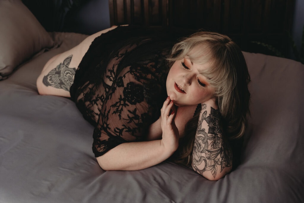 A pale, plus-size woman with blonde hair and black lace lingerie is lying on a bed, posing with her hand touching her chin and neck. She has tattoos on her arm and leg, featuring a snake, leaves, and flowers. Featured in the blog post “The Sexy Plus Size Lingerie." Sexy, plus size, plus size lingerie, lingerie for all, female empowerment, feel good, body inclusivity, comfortable, look good, lingerie, beautiful bras, plus size panties, shop inclusive, support plus size brands, boudoir lingerie, plus size boudoir. Photograph by Embodied Art Boudoir.