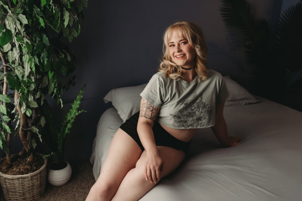 Woman sitting on the edge of a bed, wearing a gray t shirt and black shorts. Photo by Embodied Art Boudoir. Body positivity, Body positive, love yourself, love your body, body acceptance, growth, self-acceptance, women's beauty, inner beauty, goddess, challenge societal standards, redefine beauty, champion diversity, every body is beautiful, colorado boudoir, denver boudoir, boulder boudoir, colorado springs boudoir, boudoir ideas, boudoir poses, boudoir inspiration, photography inspiration
