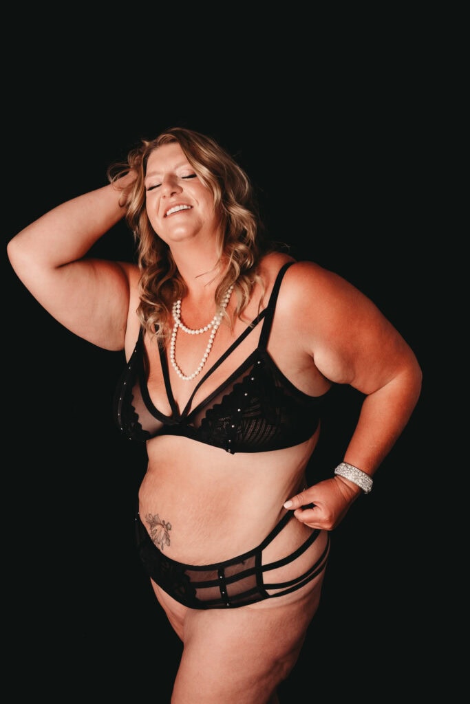Blonde women standing wearing a black bra and panties, smiling wearing a big pearl necklace. Photo by Embodied Art Boudoir. Body positivity, Body positive, love yourself, love your body, body acceptance, growth, self-acceptance, women's beauty, inner beauty, goddess, challenge societal standards, redefine beauty, champion diversity, every body is beautiful, colorado boudoir, denver boudoir, boulder boudoir, colorado springs boudoir, boudoir ideas, boudoir poses, boudoir inspiration, photography inspiration