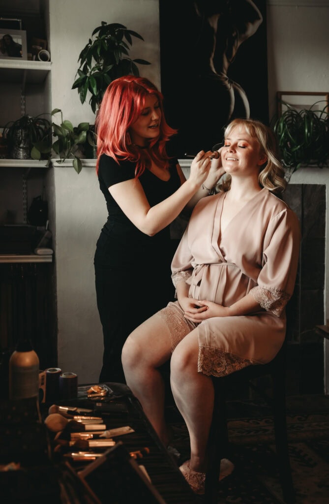 Woman with red hair applying makeup to a female client who is sitting on a stool wearing a baby pink silky robe, she has peachy eyeshadow on her eyes and a soft lip. Featured in the blog post "In Conversation With Embodied Art Boudoir's Hair & Makeup Artist" Boudoir photography, Colorado boudoir, Denver boudoir, female photographer, pampering, hair and makeup, feel good, makeup artist, interview, expanding, self love, celebrity treatment, self care, attractive, beautiful, stylish, hair styles, glamorous. Photograph by Embodied Art Boudoir,