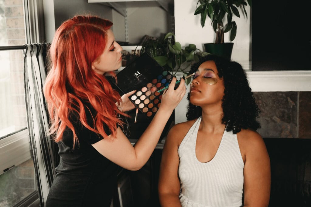 Red haired woman wearing a black t-shirt applying a purple eyeshadow to the eyes of a black female client who is sitting in the makeup chair wearing a white sleeveless tank top. Featured in the blog post "In Conversation With Embodied Art Boudoir's Hair & Makeup Artist" Boudoir photography, Colorado boudoir, Denver boudoir, female photographer, pampering, hair and makeup, feel good, makeup artist, interview, expanding, self love, celebrity treatment, self care, attractive, beautiful, stylish, hair styles, glamorous. Photograph by Embodied Art Boudoir,