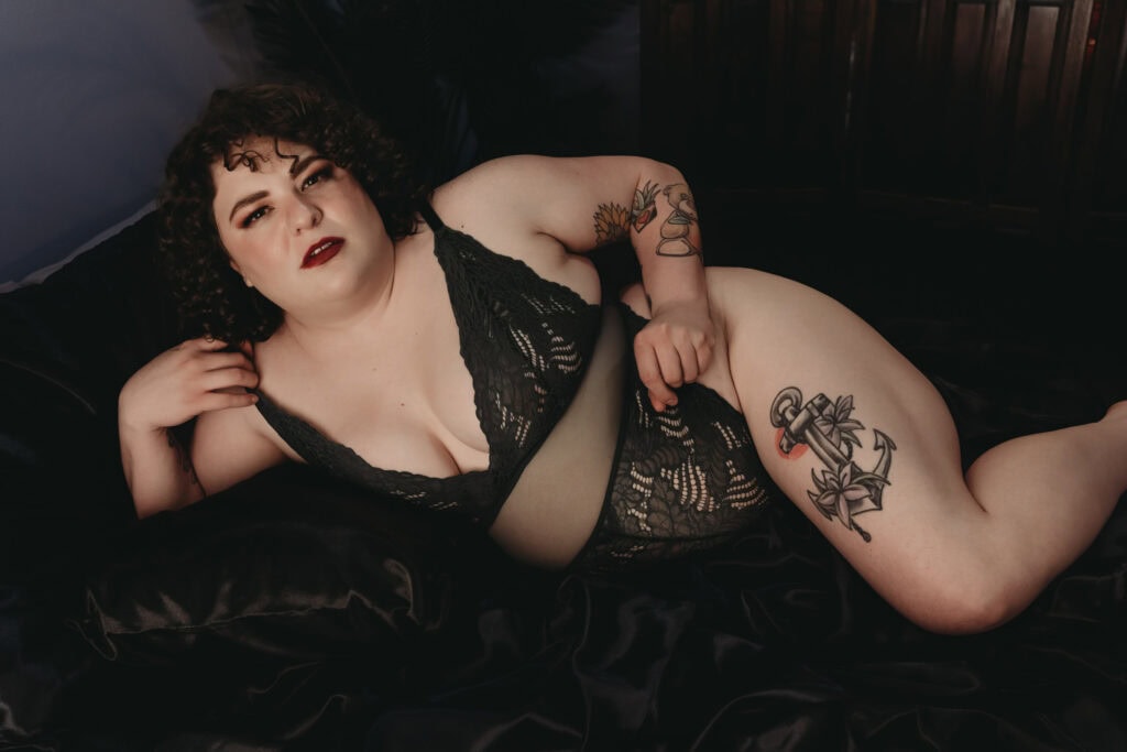 plus-sized woman with short curly bob lying on a bed posing wearing a low cut lacy body suit that is black, with a see thru slit across the stomach, she has on dark eye makeup and red lipstick, with her large tattoos on display. Featured in the blog, "Where to Buy Bras When You Have Huge Boobs." bra, bra shopping, large breasts, big boob, huge boobs, bra intervention, limited size, right fit, comfortable bra, lingerie, bra sizing system, right shape, measure yourself, bra cup, brands, colorado, denver boudoir, boudoir lingerie, boudoir outfits, boudoir bras. Photo by Embodied Art Boudoir.