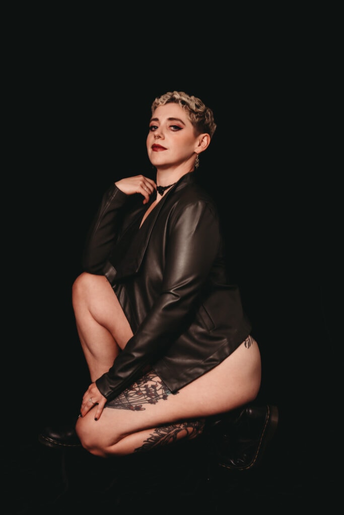 White blonde woman sitting facing the camera, wearing nothing but black army boots and a leather jacket. She looks confidently at the camera and is wearing dark eye makeup and dark red lipstick. Featured in the blog post "Femme vs Masc Boudoir Styling, Outfits, and Posing" colorado boudoir photography, Self love, Colorado boudoir photographer, gender exploration, gender neutrality, self-exploration, sex, gender, fashion, style, confidence, body image, be yourself, self-discover, self-discovery, comfortability, boudoir, boudoir posing, boudoir imagery, boudoir photo, inclusivity. Picture by Embodied Art Boudoir.