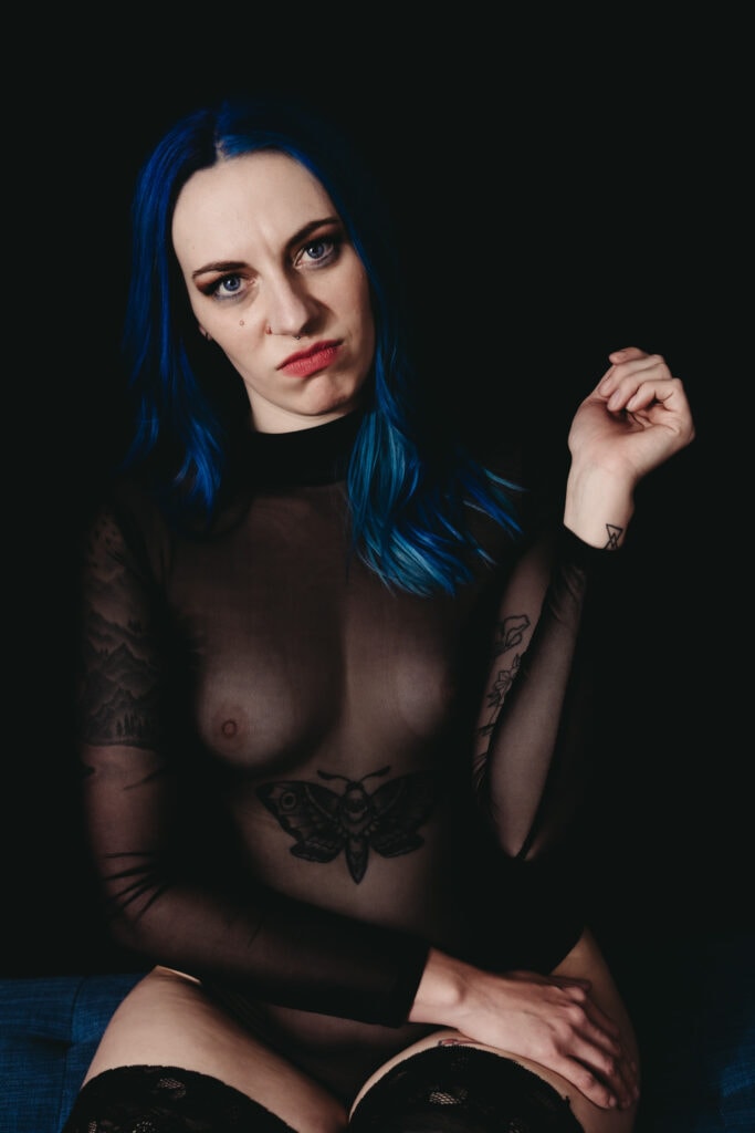 Woman with dyed blue hair looking sassily and aggressively at the camera. She has dark eye markup and dark lipstick and is wearing a see-through black body suit with knee-high socks. Featured in the blog post "Femme vs Masc Boudoir Styling, Outfits, and Posing" colorado boudoir photography, Self love, Colorado boudoir photographer, gender exploration, gender neutrality, self-exploration, sex, gender, fashion, style, confidence, body image, be yourself, self-discover, self-discovery, comfortability, boudoir, boudoir posing, boudoir imagery, boudoir photo, inclusivity. Photo by Embodied Art Boudoir.