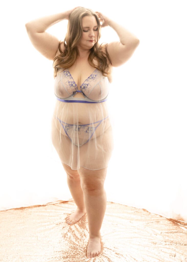 Plus sized mature woman standing up with her hands posed gracefully on her curly brown long hair. She is wearing a long chemise that is see through with a purple and white g string and bra. Featured in the blog post "Femme vs Masc Boudoir Styling, Outfits, and Posing" colorado boudoir photography, Self love, Colorado boudoir photographer, gender exploration, gender neutrality, self-exploration, sex, gender, fashion, style, confidence, body image, be yourself, self-discover, self-discovery, comfortability, boudoir, boudoir posing, boudoir imagery, boudoir photo, inclusivity. Photograph by Embodied Art Boudoir.