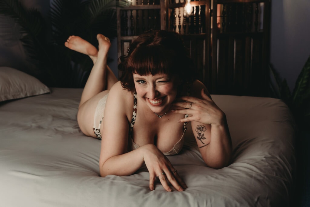 Woman with short red hair with bangs facing the camera winking at it, wearing a black and white bra and matching panties. She is lying on the bed with white sheets. Featured in the blog post "Femme vs Masc Boudoir Styling, Outfits, and Posing" colorado boudoir photography, Self love, Colorado boudoir photographer, gender exploration, gender neutrality, self-exploration, sex, gender, fashion, style, confidence, body image, be yourself, self-discover, self-discovery, comfortability, boudoir, boudoir posing, boudoir imagery, boudoir photo, inclusivity. Image by Embodied Art Boudoir.