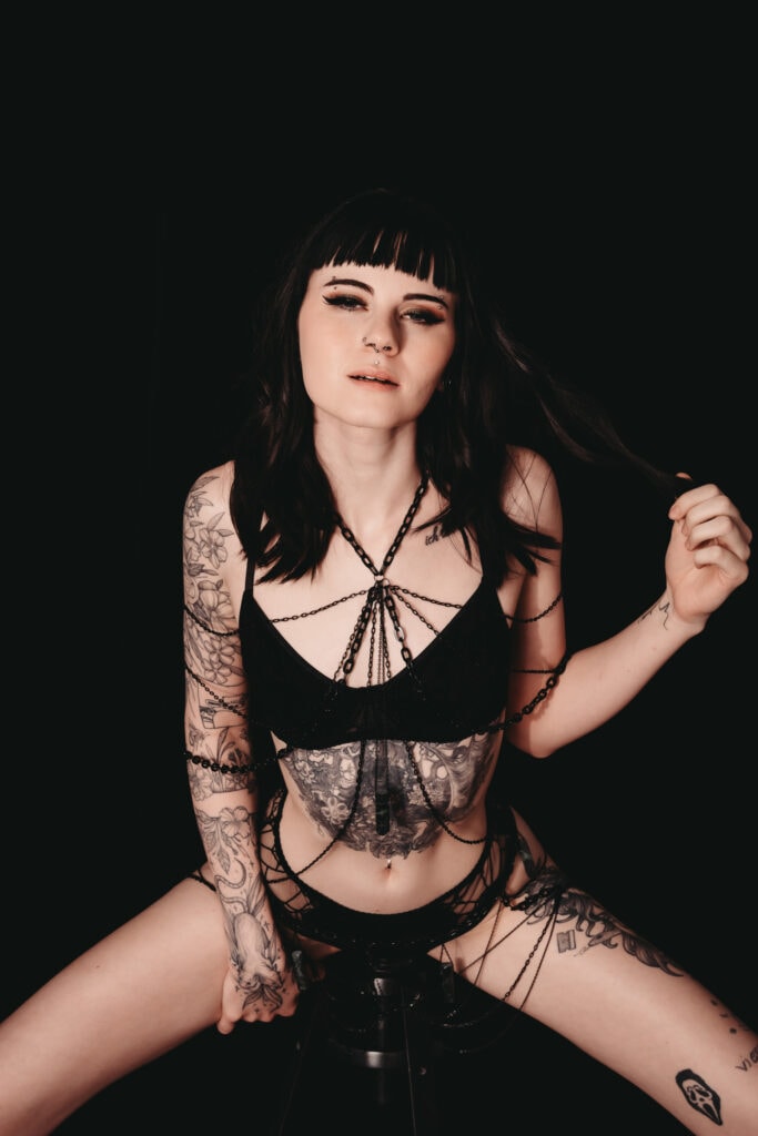 Heavily tattooed young woman with jet-black hair is sitting on a stool with her legs open, looking at the camera seriously and seductively. She is wearing a matching black lingerie set that showcases her black tattoos. Featured in the blog post "Femme vs Masc Boudoir Styling, Outfits, and Posing" colorado boudoir photography, Self love, Colorado boudoir photographer, gender exploration, gender neutrality, self-exploration, sex, gender, fashion, style, confidence, body image, be yourself, self-discover, self-discovery, comfortability, boudoir, boudoir posing, boudoir imagery, boudoir photo, inclusivity. Image by Embodied Art Boudoir.