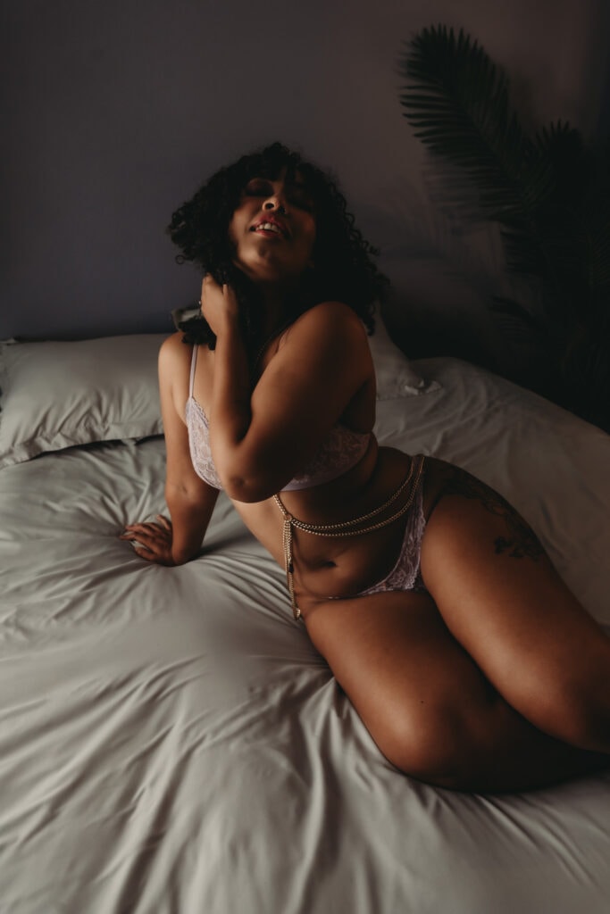 Young black woman with short curly hair sitting on bed with her hair thrown back confidently wearing a matching purple bra and panties with a silver body chain. She is sat confidently atop a bed with grey sheets. Featured in the blog post "Femme vs Masc Boudoir Styling, Outfits, and Posing" colorado boudoir photography, Self love, Colorado boudoir photographer, gender exploration, gender neutrality, self-exploration, sex, gender, fashion, style, confidence, body image, be yourself, self-discover, self-discovery, comfortability, boudoir, boudoir posing, boudoir imagery, boudoir photo, inclusivity. Image by Embodied Art Boudoir.