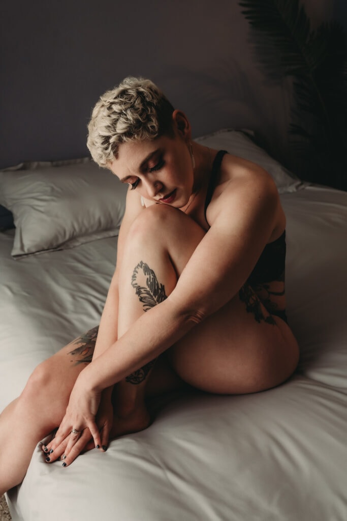 Blonde woman with short pixie cut hair shielding her body by hugging her knee to her chest. She looks solemn and calm. Featured in the blog post "Femme vs Masc Boudoir Styling, Outfits, and Posing" colorado boudoir photography, Self love, Colorado boudoir photographer, gender exploration, gender neutrality, self-exploration, sex, gender, fashion, style, confidence, body image, be yourself, self-discover, self-discovery, comfortability, boudoir, boudoir posing, boudoir imagery, boudoir photo, inclusivity. Image by Embodied Art Boudoir.