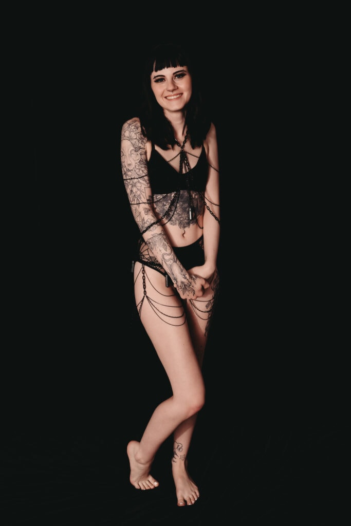 Black black-haired woman covered with black tattoos stands coyly facing the camera, wearing a black matching bra and underwear with a body chain that is covering her chest and legs. Featured in the blog post "Femme vs Masc Boudoir Styling, Outfits, and Posing" colorado boudoir photography, Self love, Colorado boudoir photographer, gender exploration, gender neutrality, self-exploration, sex, gender, fashion, style, confidence, body image, be yourself, self-discover, self-discovery, comfortability, boudoir, boudoir posing, boudoir imagery, boudoir photo, inclusivity. Photography by Embodied Art Boudoir.