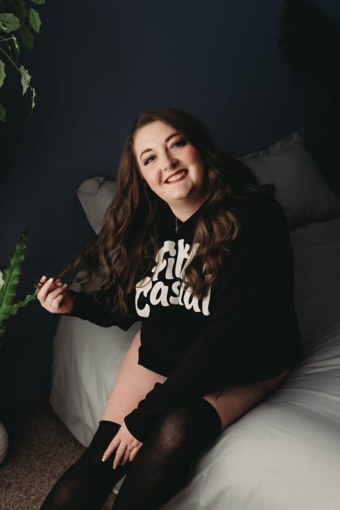 Young brunette woman with curly hair smiling at camera while she play with her hair, wearing a black hoodie and black thigh high leggings. Featured in the blog post "Femme vs Masc Boudoir Styling, Outfits, and Posing" colorado boudoir photography, Self love, Colorado boudoir photographer, gender exploration, gender neutrality, self-exploration, sex, gender, fashion, style, confidence, body image, be yourself, self-discover, self-discovery, comfortability, boudoir, boudoir posing, boudoir imagery, boudoir photo, inclusivity. Image by Embodied Art Boudoir.